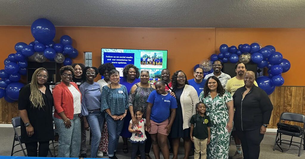 A large group of adults and children pose together for a photo inside a community room decorated for an event. The group stands shoulder to shoulder in front of an orange wall, framed on both sides by blue balloon arches with a few gold-accented balloons. Several people are smiling at the camera, and the group includes community members of different ages, including two young children near the front. Behind the group, a screen displays a slide encouraging attendees to follow the organization on social media and sign up for a newsletter, with small event photos shown on the slide. The room has fluorescent ceiling lights, folding chairs, wood paneling along the lower walls, and blue tablecloths on tables in the foreground. On the tables are cups, plates, notebooks, a pen, a small orange, and an orange folder. The overall atmosphere feels celebratory, communal, and informal, suggesting a community gathering, workshop, or program celebration.